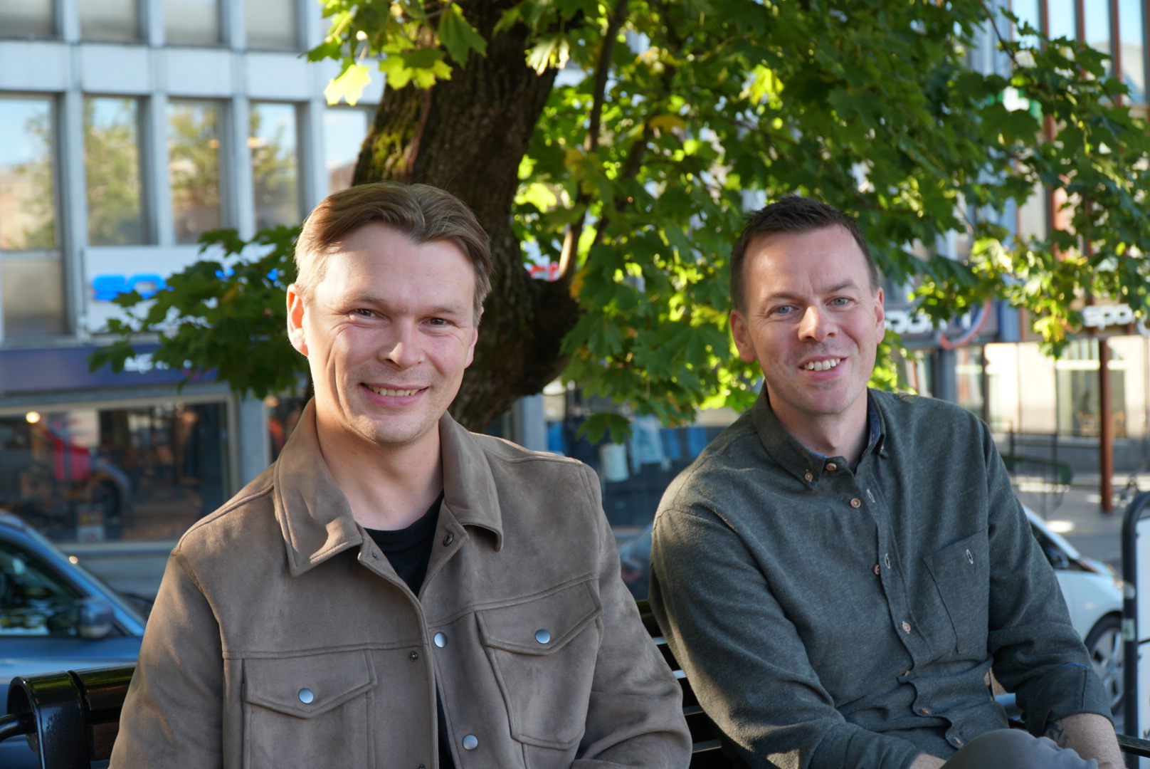 Bjarte Solhaug og Alexander Henningsen sitter smilende på en benk under et lønnetre i et solfylt bysentrum.
